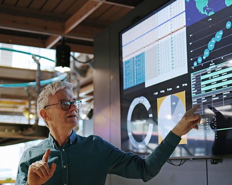 Man conducting a presentation with the aid of a large screen. The screen is displaying graphs and data
