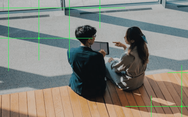 From a high angle view, two businesspersons are seated on chairs in the lobby. A green grid is overlaid on the image
