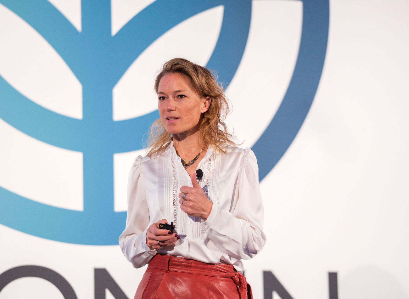 A woman in a white blouse speaking on stage with a microphone, holding a presentation clicker, standing in front of a large event logo.