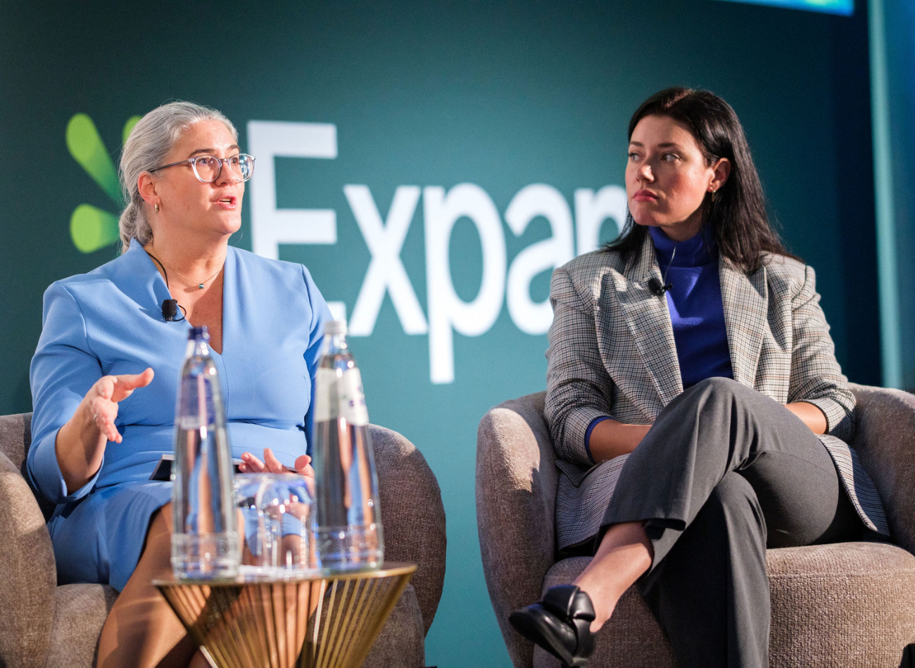 Two women seated on stage during a panel discussion; one in a light blue outfit speaking with hand gestures, the other in a grey blazer listening attentively, with the “Expana” logo in the background.