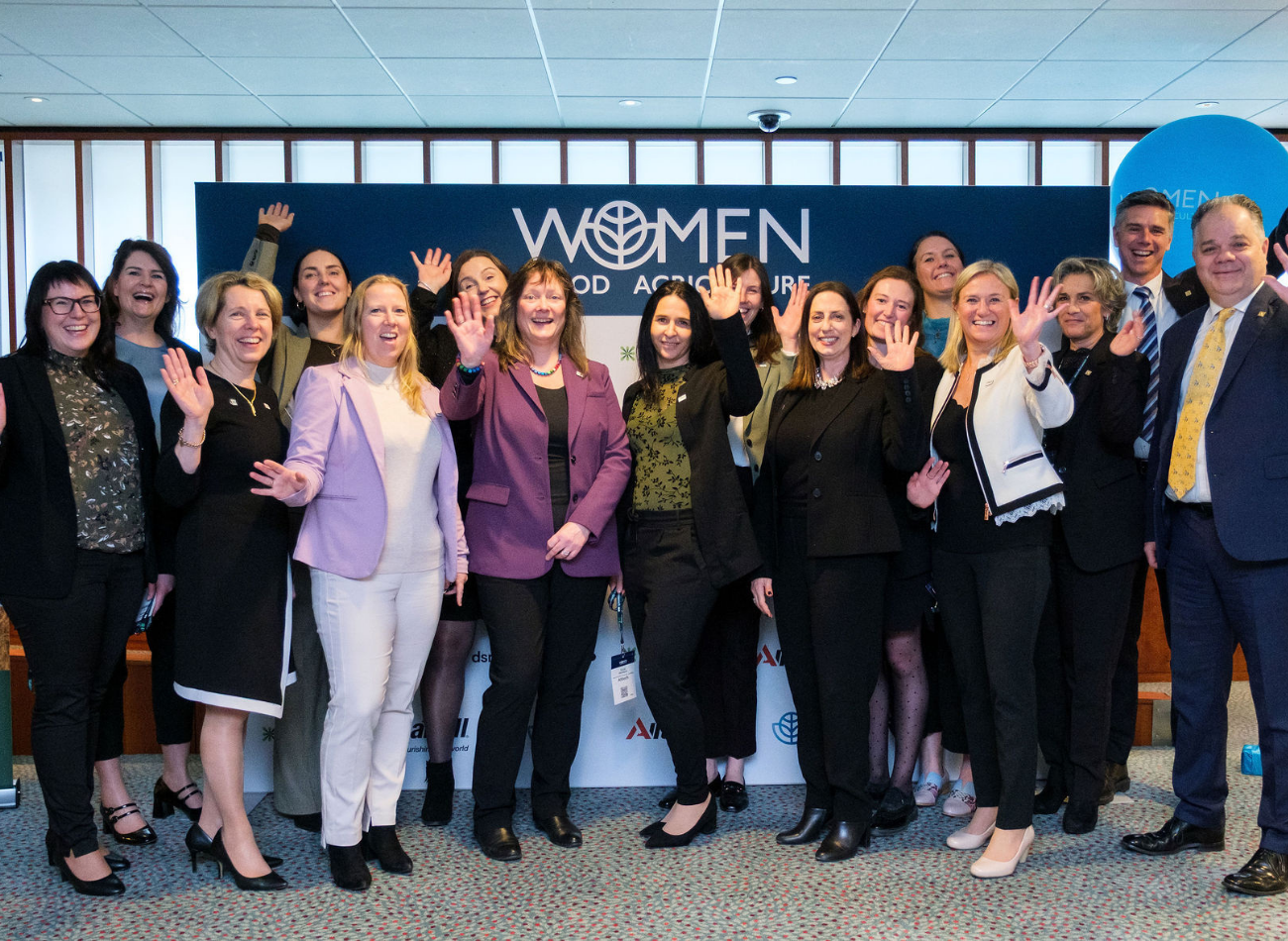 A group of professionally dressed women and a few men smiling and waving at the camera while standing in front of a “Women in Food & Agriculture” event backdrop.