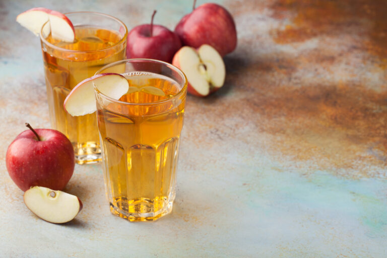 Two glasses of apple juice garnished with fresh red apple slices, surrounded by whole and sliced apples.