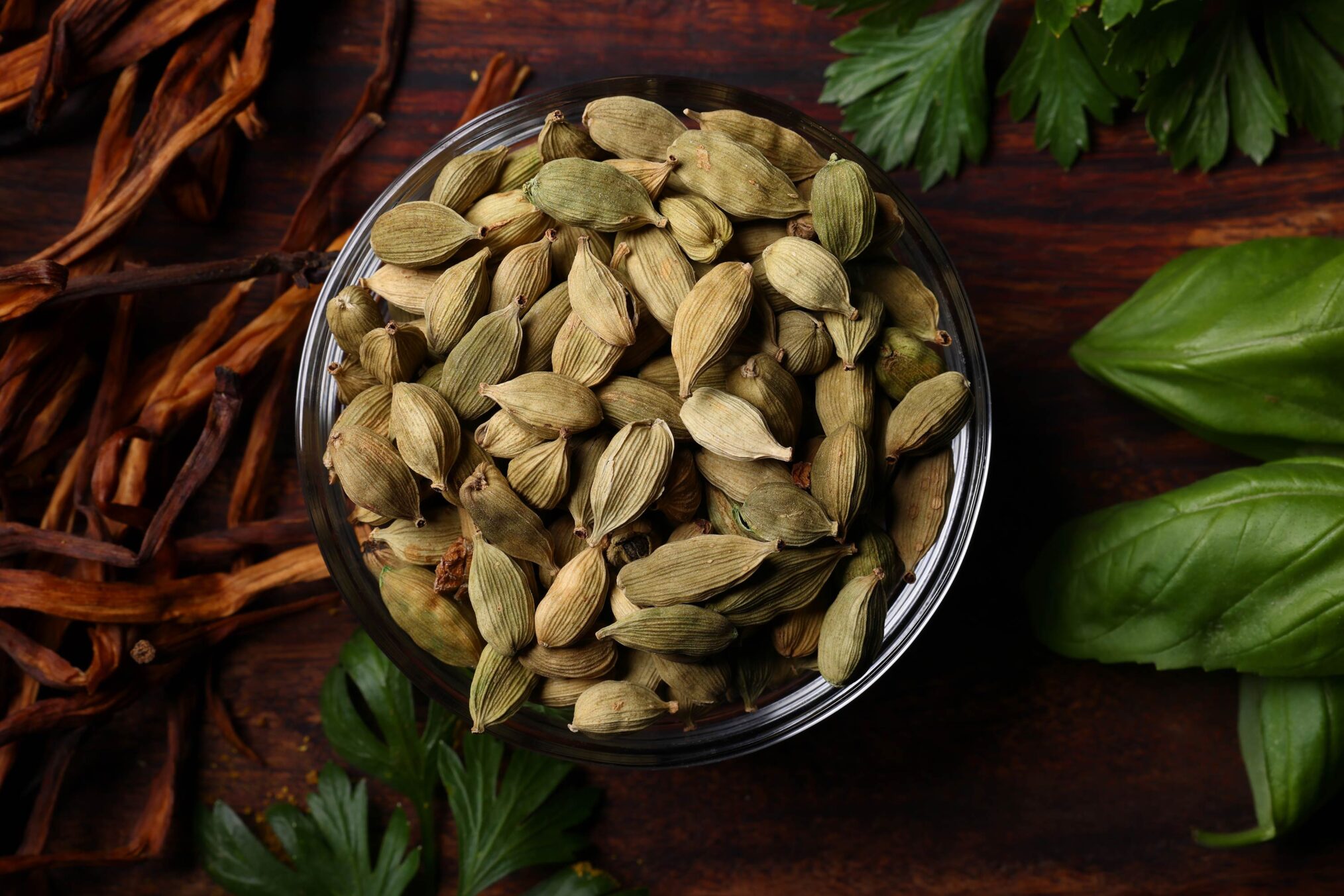 Whole green cardamom pods in a glass bowl surrounded by herbs and spices